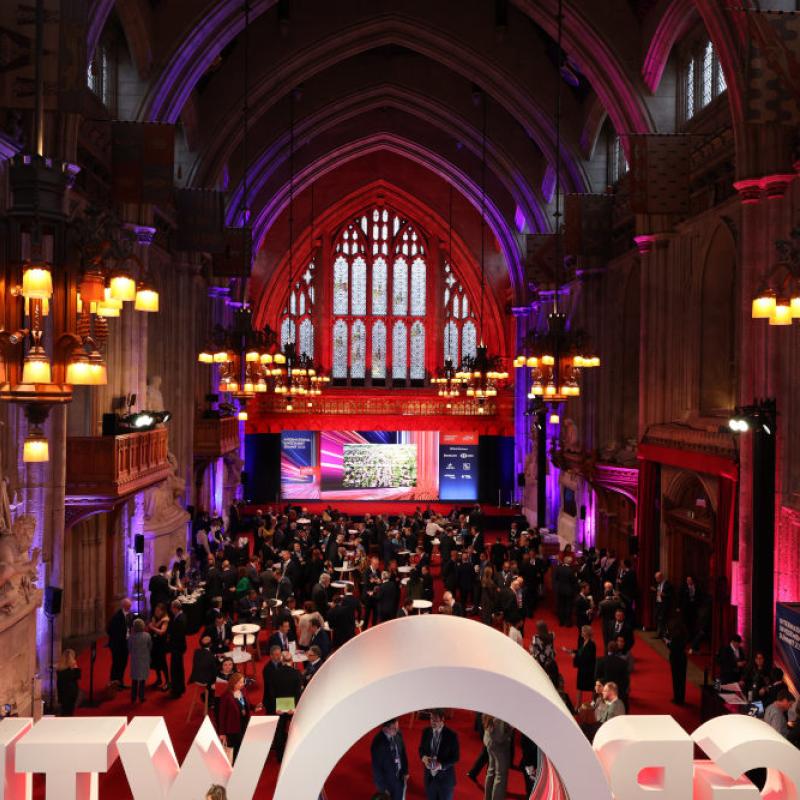 Grand hall with high ceilings hosting a formal event with attendees, stage, and large illuminated signage.