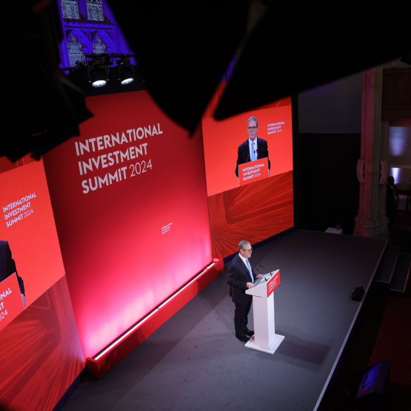 A speaker in a suit delivers a speech at the International Investment Summit 2024, standing at a podium on a red-lit stage with large screens.