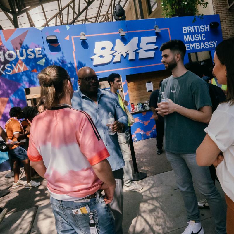 A group of people converses outside the British Music Embassy booth at an outdoor event, with a colourful backdrop and lively atmosphere.