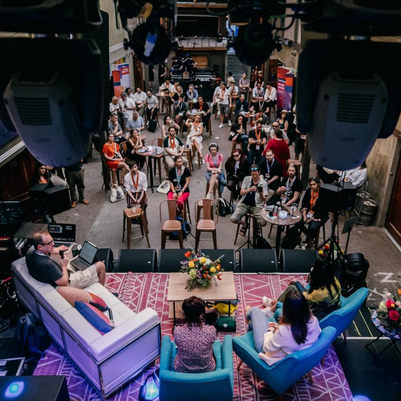 A panel discussion takes place on a stage with three speakers seated, while an engaged audience listens in a casual, intimate venue setting.