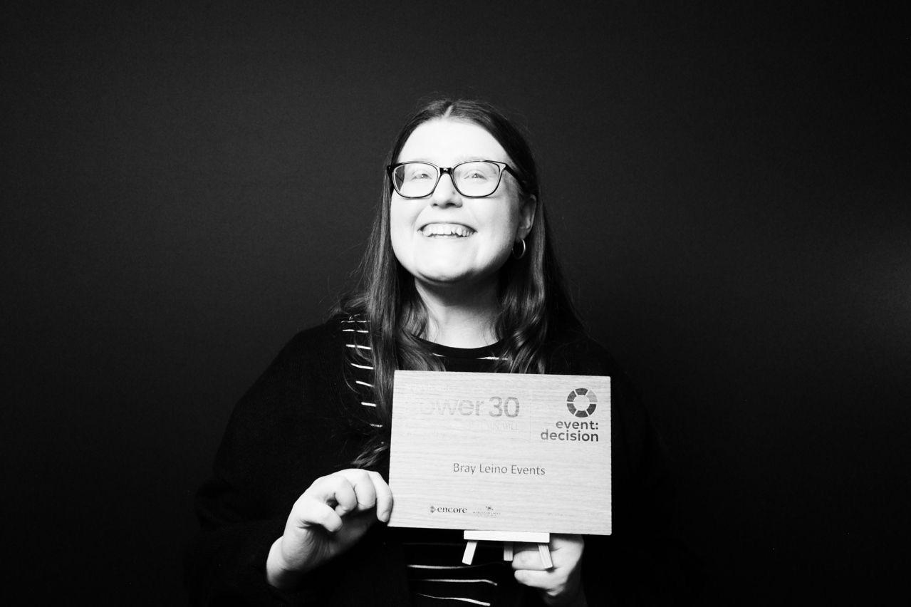 A smiling person with glasses holds a wooden award plaque that reads "Power 30" and "Bray Leino Events" against a dark background.