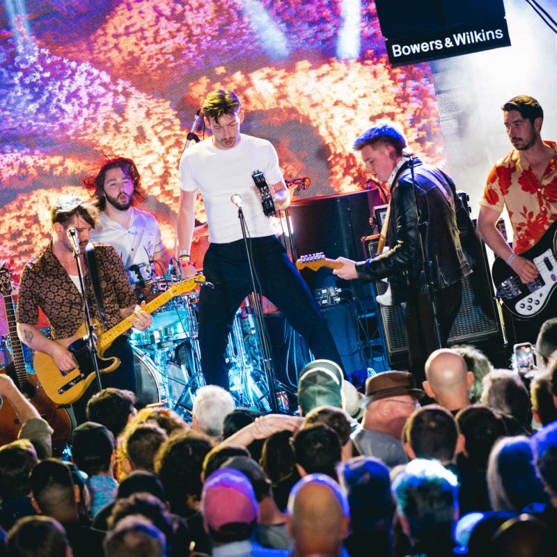 A live band performs on stage with energetic lighting, as a crowd watches. The lead singer holds a tambourine while musicians play guitars.